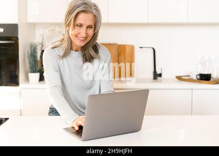 Positivo anziano donna seduta relax in cucina, lavoro su laptop, sorridente, moderna matura donna 60 godere di tempo libero week-end a casa, navigazione, web surf su computer, shopping, digitando messaggio Foto Stock