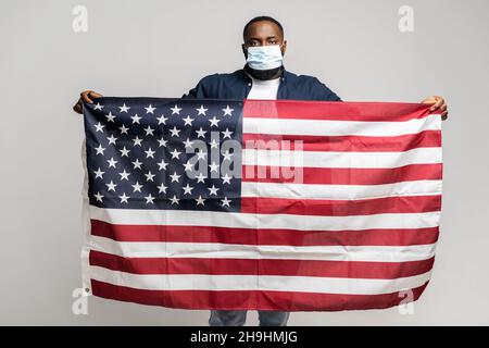Grave uomo afroamericano in maschera medica protettiva in piedi e guardando la macchina fotografica, maschio tenendo bandiera USA sparso in braccia di fronte a lui, isolato su sfondo grigio Foto Stock