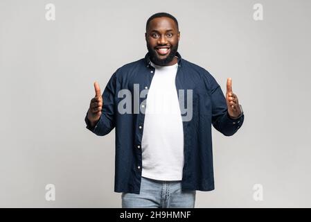 L'uomo africano positivo con la barba guarda la macchina fotografica, tenendo due mani davanti a lui mostrando le dimensioni su sfondo grigio isolato in studio, indossando una camicia casual, sorridendo gioiosamente, presentando qualcosa Foto Stock