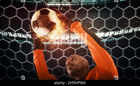 Primo piano di una scena calcistica durante la partita notturna con un portiere che cerca di catturare una palla ardente Foto Stock