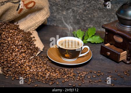 Kaffee Komposition mit dampfender Kaffeetasse, Kaffeebohnen in Kaffeesack, historischer Kaffeemühle und Kaffeeblätter auf einem Holztisch Foto Stock