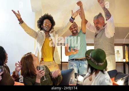 gruppo multietnico di uomini che si acclamano per qualcosa guardando lo smartphone, mentre gruppo di donne stupite che li guardano, festa di festa e sport Foto Stock