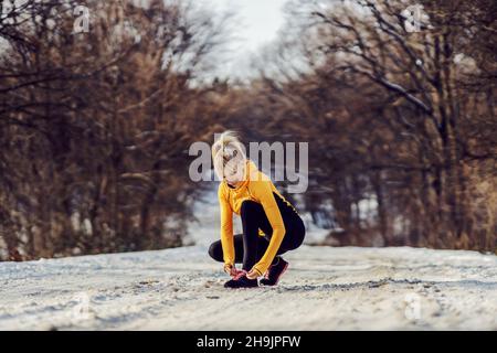 Sport swoman accovacciato su sentiero nevoso in natura in inverno e legando la sua scarpa. Sportswear, fitness invernale, vita sana Foto Stock