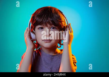 Ritratto di ragazzino, bambino che ascolta la musica in cuffie isolate su sfondo blu studio in luce al neon, filtro. Foto Stock