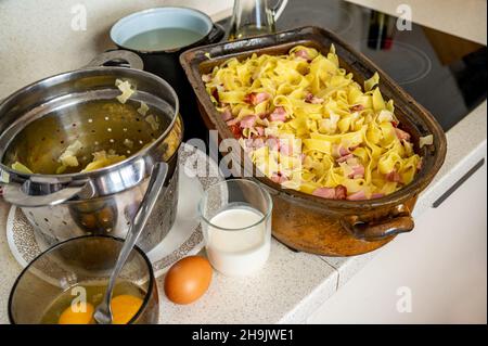 Teglia da forno in terracotta con pasta e carne affumicata, latte, uova, pentola e colino sul piano cottura. Preparazione del pasto in cucina. Foto Stock