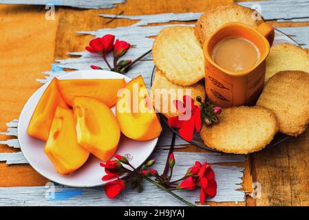 Frutta kaki affetta sul piatto, biscotto al burro, tazza di caffè all'arancia e fiore rosso sul tavolo di legno graffiato. Dolce cibo aranciato. Foto Stock