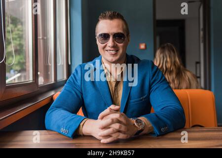 Un giovane uomo d'affari sorridente di aspetto europeo elegante ritratto con occhiali si siede a un tavolo in un caffè a guardare. Foto Stock