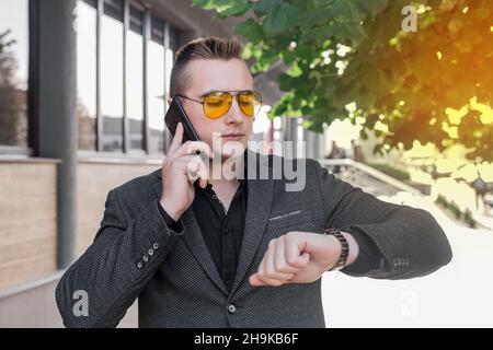 Un giovane ragazzo serio, uomo d'affari di ritratto europeo in occhiali da sole parla su un telefono cellulare o smartphone e guarda il tempo su una mano w Foto Stock