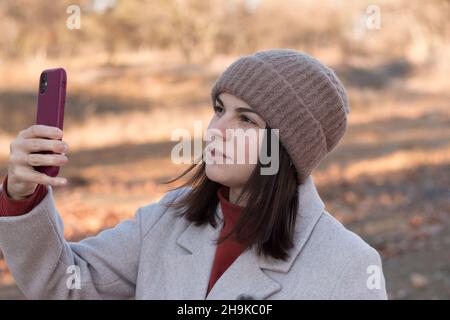 Una giovane ragazza in un cappello e cappotto in maglia tiene un telefono nelle sue mani sullo sfondo di un paesaggio di parco autunno Foto Stock