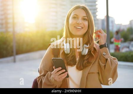 La giovane donna che ride guarda la macchina fotografica nei vestiti autunnali scegliendo la playlist sullo smartphone all'aperto al tramonto Foto Stock