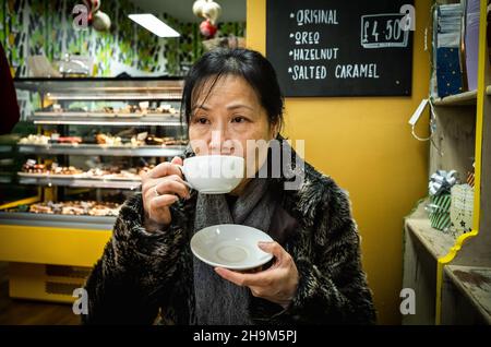 Una donna vietnamita che indossa un cappotto e una sciarpa in pelliccia sintetica si siede in un caffè artigianale al cioccolato chiamato Cenu cacao nella King's Parade, Cambridge, Regno Unito Foto Stock