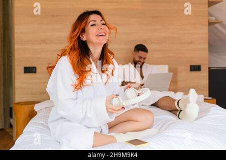 Donna con cuffie ride seduto a gambe incrociate sul letto. Donna dai capelli rossi in un accappatoio con cuffie. Foto Stock