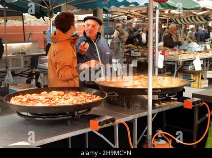 SAINT JEAN DE LUZ, FRANCIA - 24 maggio 2019: Uomo con un cappello basco in discussione con un uomo che prepara la paella all'aria aperta Foto Stock