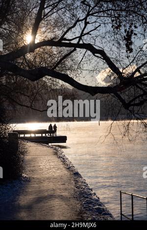 Persone che camminano su un percorso a piedi nella baia di Stoccolma in una gelida giornata invernale con acqua fumante, albero di betulla di fronte e accogliente luce solare. Ora invernale Foto Stock
