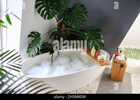 Vasca da bagno piena di bolle decorata con piante tropicali monstera. Prodotti per la cura del corpo ecologici su scaffali in legno Foto Stock
