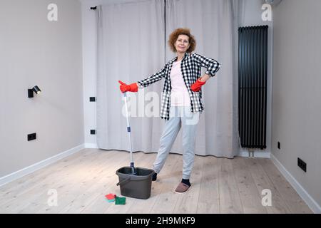 Persone, lavori domestici e concetto di pulizia. Donna anziana felice in guanti protettivi pulisce il pavimento e danze a casa in appartamento vuoto prima di muoversi Foto Stock