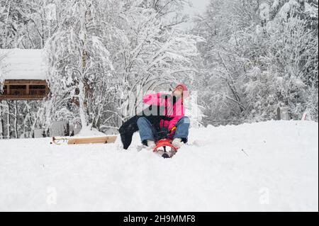 Ragazza allegra felice su una slade con il suo cane nero labrador che salta amorevolmente sul suo esterno in una bella natura invernale nevosa. Foto Stock