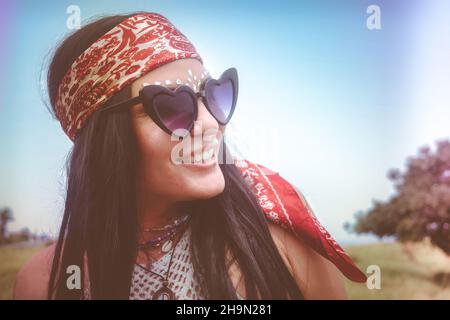 Ritratto in stile retrò opaco di una donna bohémien, con capelli neri lisci lunghi, bandana vintage, gioielli con perline, divertenti occhiali da sole, glitter. Foto Stock
