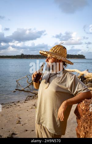 Uomo giovane con cappello e bottiglia di birra ambrata sul bordo del lago. Concetto di vita rilassato in vacanza. Foto Stock