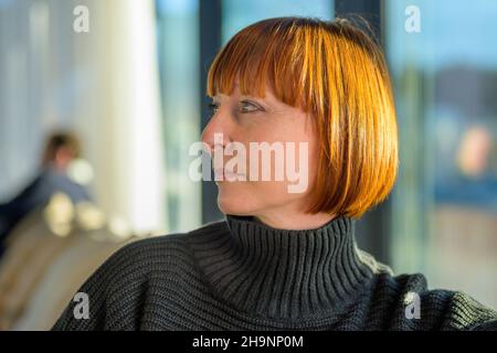 Ritratto di un attraente trendy di mezza età rosso capelli donna seduta al chiuso guardando a sinistra della cornice in una vista laterale in primo piano Foto Stock