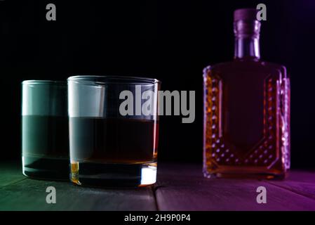 Due bicchieri con alcol e una bottiglia su un tavolo di legno in una stanza buia. Foto Stock