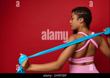 Vista dal dorso a una donna nera positiva per il corpo in abbigliamento sportivo rosa, esercizi con cinturino elastico blu, braccia tese contro lo schienale rosso Foto Stock