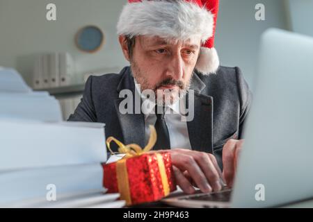 Uomo d'affari serio con cappello Babbo Natale digitando sulla tastiera del computer portatile durante la stagione natalizia nel suo ufficio, fuoco selettivo Foto Stock