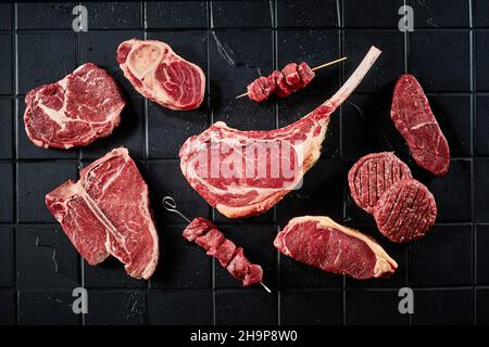 Vista dall'alto delle cotolette di hamburger tomahawk non cotte e delle bistecche di lombata con kebab shish posto su un tavolo piastrellato nero Foto Stock