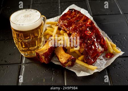 Da sopra di barattolo di birra posto vicino patate fritte yummy e costolette con salsa sul tavolo nero Foto Stock