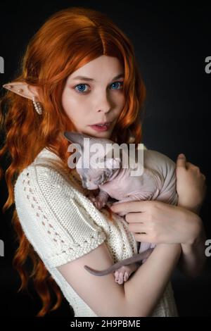 Ritratto di giovane donna coscrayer elf, abbracciando Sphinx kitten. Elf in abito in maglia bianca, con capelli ricci rossi lunghi di lusso e orecchie traforate Foto Stock