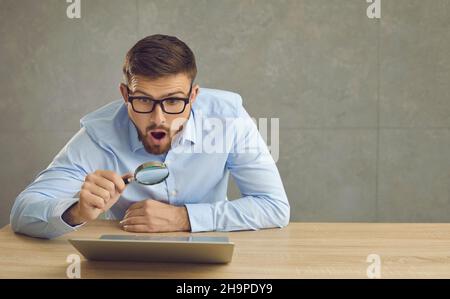 L'uomo con un'espressione sorpresa guarda lo schermo del laptop attraverso una lente d'ingrandimento. Foto Stock