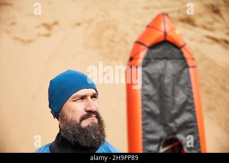 Ritratto di uomo vecchio con barba nera e pagaia in giacca blu vicino a barca sportiva arancione zattera su sfondo di dune di sabbia Foto Stock