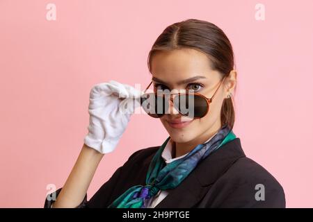 Giovane ragazza di stile, assistente di volo femminile indossando occhiali da sole guardando la macchina fotografica isolato su sfondo rosa studio. Foto Stock