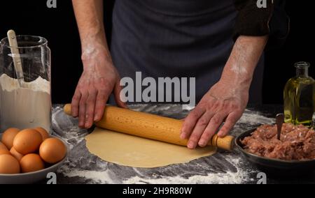 Un uomo arrotola l'impasto con un matterello per un piatto tradizionale russo gnocchi, ingredienti per l'impasto e carne macinata sul tavolo Foto Stock