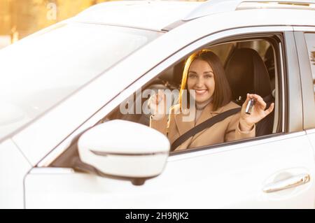 Eccitata donna allegra mostrando chiave da nuova auto, seduta all'interno guarda la fotocamera e sorridendo, urlando felicemente, donna di successo che acquista un'auto, raggiungere un concetto di obiettivo Foto Stock