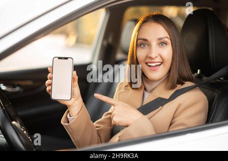 Giovane bella donna sorpresa alla guida di un'auto, la donna punta a schermo del telefono in bianco e sorride. App mobili per il driver. Vista esterna Foto Stock