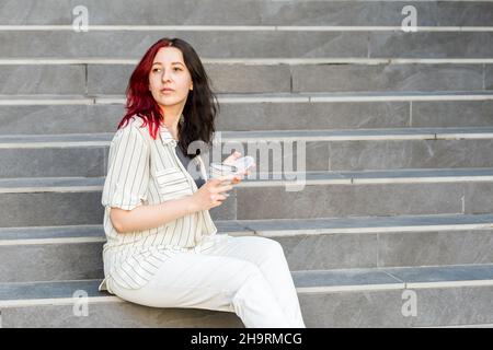 Giovane donna seduta sulle scale all'aperto, con una tazza in silicone pieghevole e un bicchiere riutilizzabile da caffè a go. Foto Stock