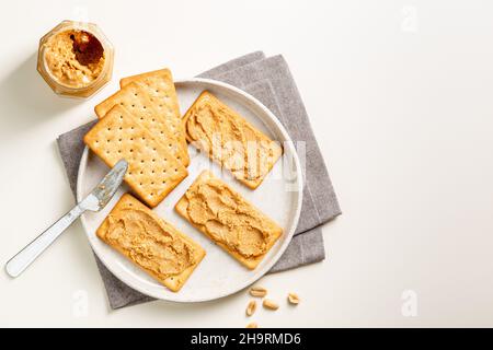 Burro di arachidi sparso su cracker di soda sul piatto visto dall'alto con coltello e vaso. Vista dall'alto Foto Stock