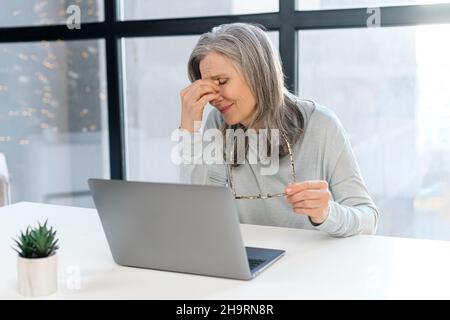 Una donna anziana, stanca e stanca, con capelli grigi, si siede con gli occhi chiusi davanti al laptop, si appoggia la testa sulla mano, si sente mal di testa e burnout Foto Stock