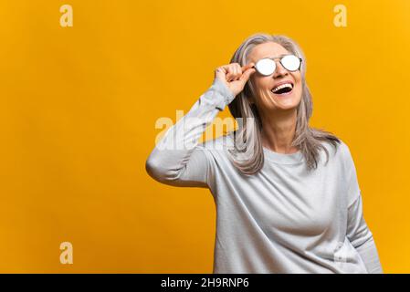 Affascinante donna anziana in occhiali da sole ridendo toothy, elegante spensierata matura donna grigio-capelli moderna facendo guardare il sole, godendo luce solare isolata su sfondo giallo Foto Stock