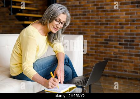 Donna matura concentrata di mezza età dai capelli grigi in occhiali utilizzando un notebook per lo studio online, seduto su un divano in salotto di fronte al computer portatile e prendere appunti, scrivere Foto Stock