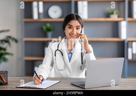 Sorridente donna indù medico terapeuta in cappotto bianco consulta paziente, parlando per telefono in clinica ufficio Foto Stock