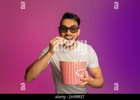 Ritratto di felice ragazzo arabo giovane in 3D bicchieri mangiare popcorn dal secchio, guardare film al cinema, neon luce Foto Stock