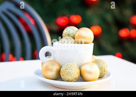 Immagine orizzontale della tazza di caffè con palle di Natale dorate all'interno e intorno. Coppia di caffè su angolo tavolo bianco, sedia grigia, albero di nuovo anno con rosso Foto Stock