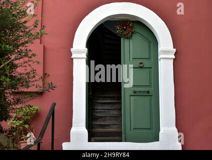 Porta in legno verde cipresso ad arco con finiture bianche decorata con una corona natalizia contro una parete di stucco rosso veneziano a Nafplio, in Grecia. Foto Stock