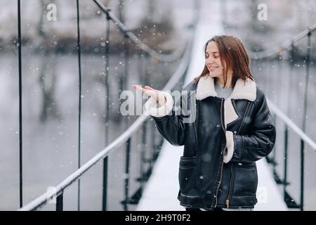 Una donna cammina e cattura fiocchi di neve sul fiume su un ponte sospeso nella giornata invernale. Giovane ragazza in abiti caldi si erge su una passerella in legno Foto Stock