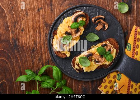 Uova strapazzate con funghi fritti e basilico sul pane su sfondo nero tavolo. Colazione fatta in casa o pranzo con uova strapazzate e funghi Foto Stock