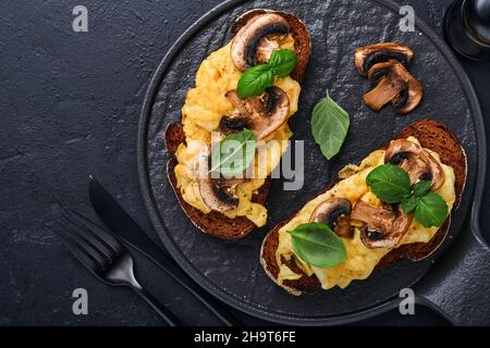 Uova strapazzate con funghi fritti e basilico sul pane su sfondo nero tavolo. Colazione fatta in casa o pranzo con uova strapazzate e funghi Foto Stock