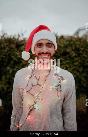L'uomo di razza mista ride in santa Hat e maglione indossando le luci di vacanza intorno al collo in inverno fuori di fronte all'albero sempreverde Foto Stock