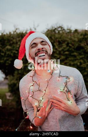 L'uomo di razza mista ride in santa Hat e maglione indossando le luci di vacanza intorno al collo in inverno fuori di fronte all'albero sempreverde Foto Stock